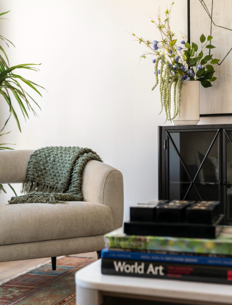 A modern, luxury living room with a plush chair and blanket, a credenza with exotic flowers, and modern art books on a table