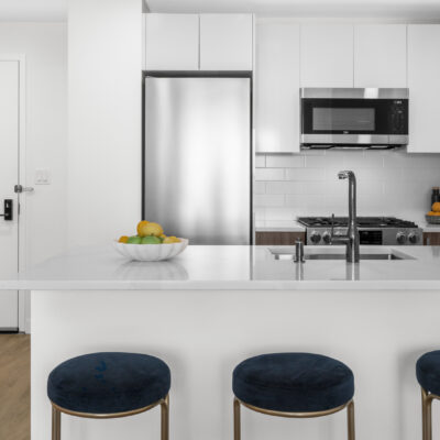 A modern kitchen with white marble counters, stools seated at an island, stainless steel appliances, and a bowl of fruit