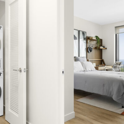 Stacked, high efficiency washer and dryer in an open closet next to a bedroom with large windows featuring city skyline views