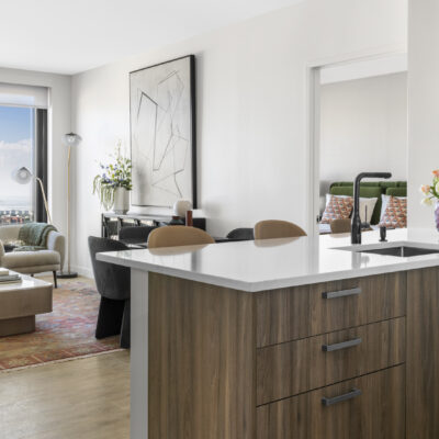 A modern, open concept kitchen with white marble counters overlooking a sunlit living room with floor to ceiling windows