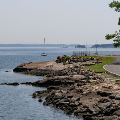 A pedestrian walkway along the New York coastline in New Rochelle with boats in the water and park benches along the path